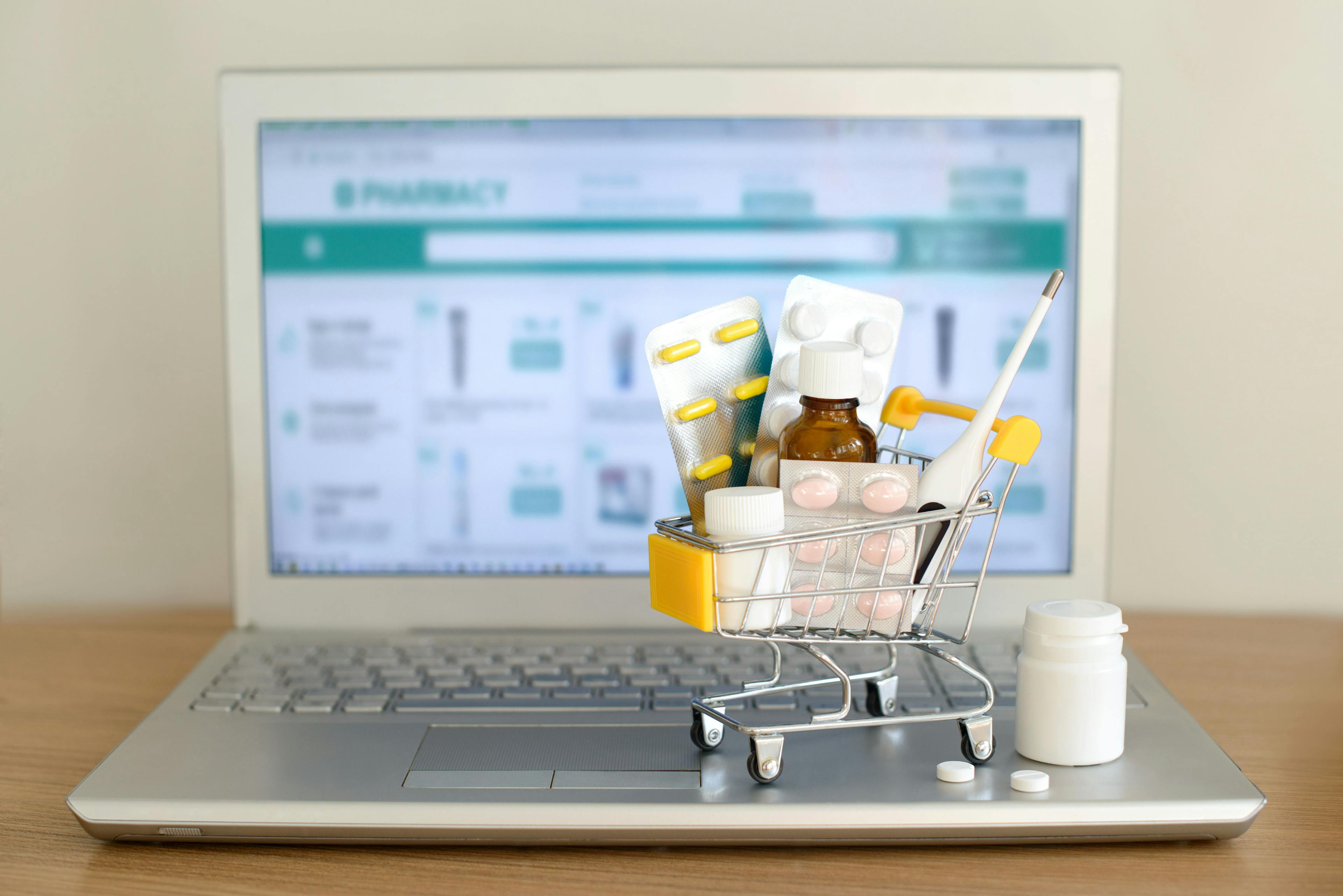 Shopping cart toy with medicaments in front of laptop screen with pharmacy web site on it. Pills, blister packs, medical bottles, thermometer set. Health care and internet shopping. Image Credit: Adobe Stock Images/evso