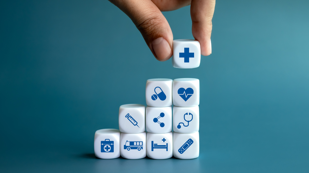 Healthcare medical, wellness plan and insurance concept. Health, care and medical element icon symbols on clean white blocks stacking as a graph arranged by doctor's hand on blue background. Image Credit: Adobe Stock Images/tete_escape