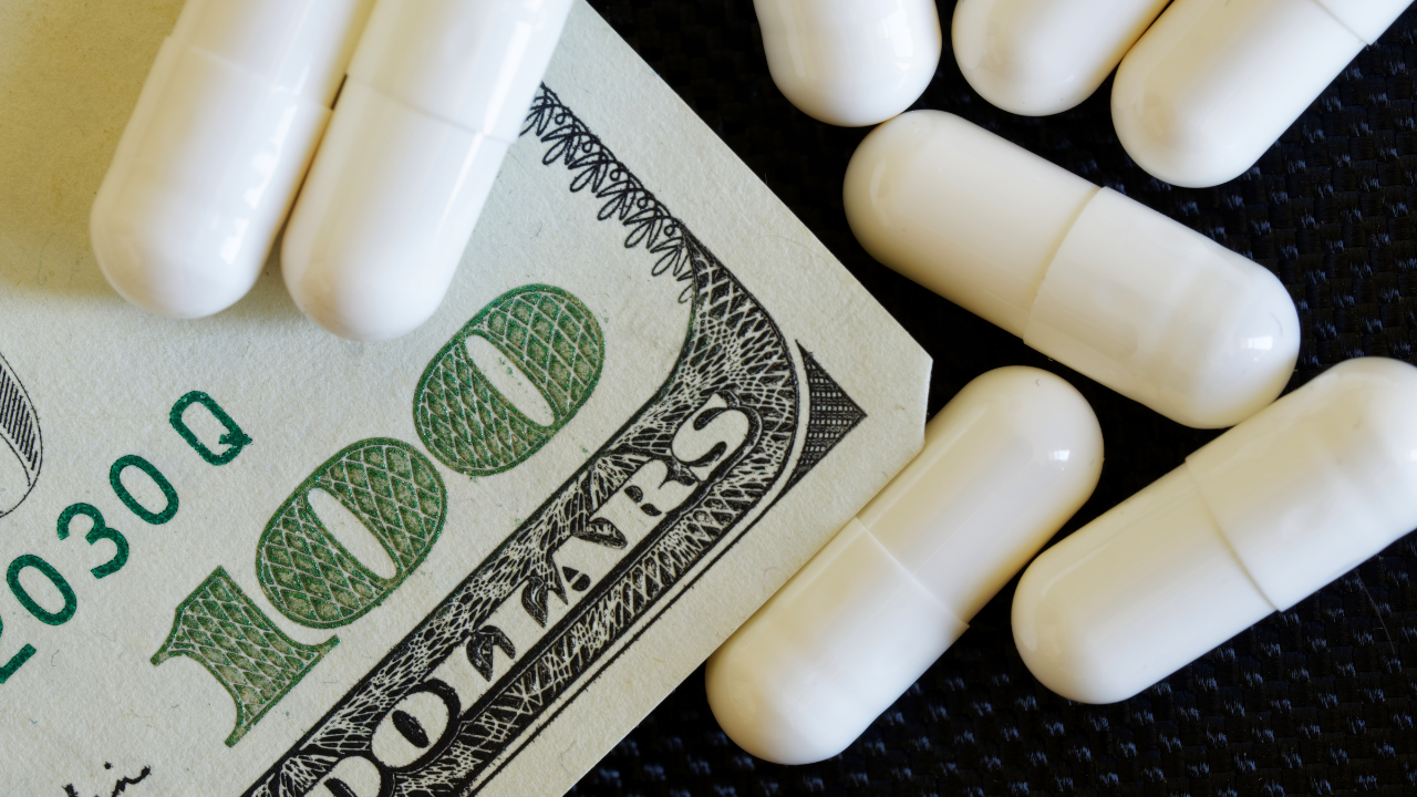 A hundred-dollar bill lies next to white capsule tablets. The problem is the high price of medicines and health insurance. Image Credit: Adobe Stock Images/lexp880