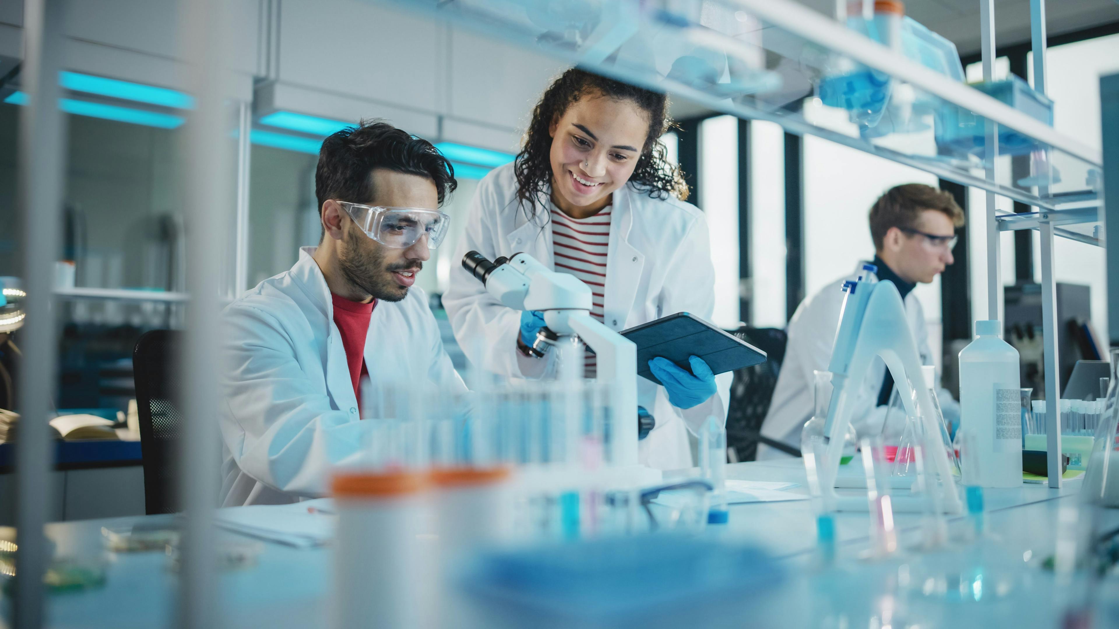Modern Medical Research Laboratory: Portrait of Latin and Black Young Scientists Using Microscope, Digital Tablet, Doing Sample Analysis, Talking. Diverse Team of Specialists work in Advanced Lab. Image Credit: Adobe Stock Images/Gorodenkoff