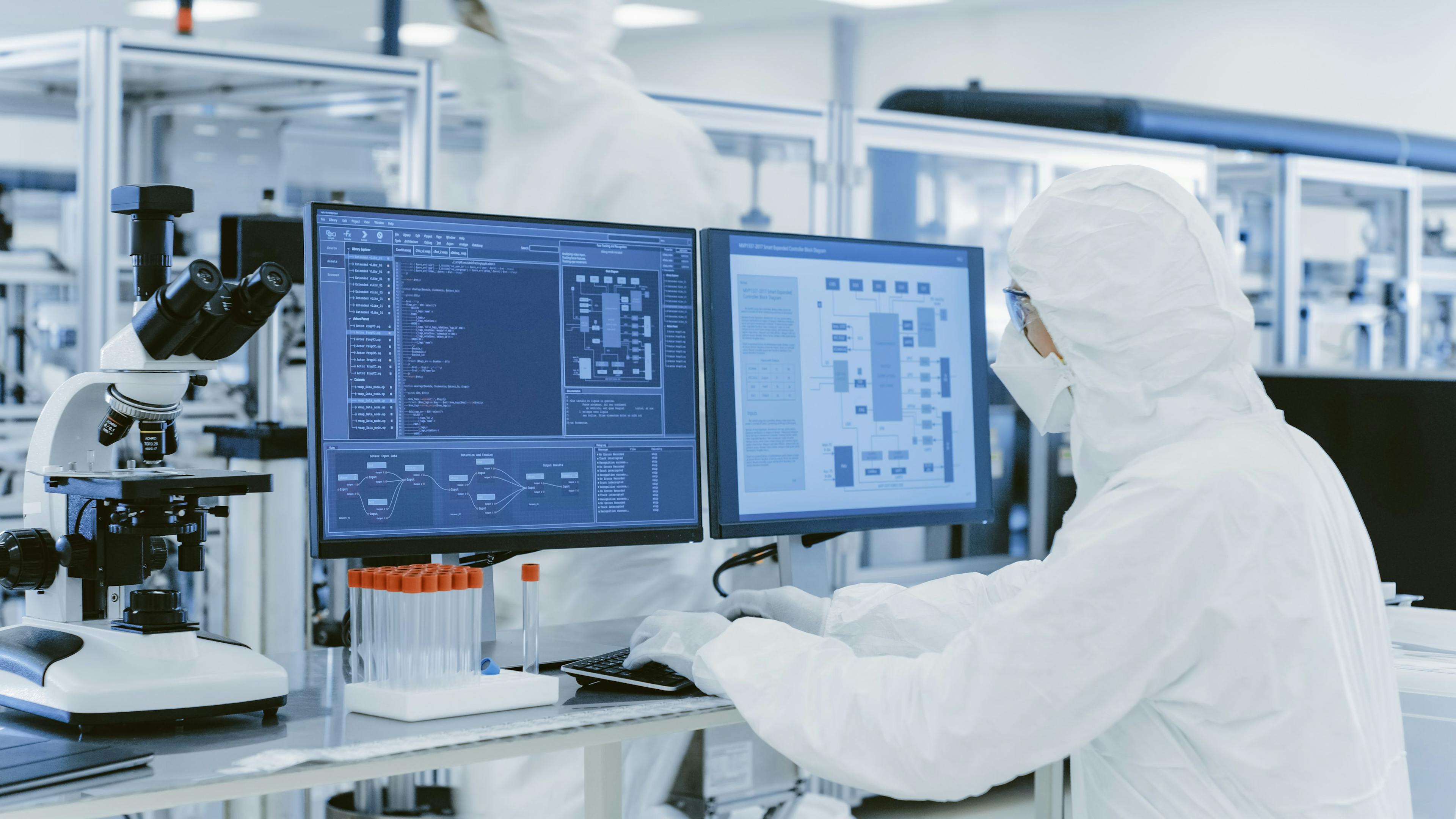 In Laboratory Over the Shoulder View of Scientist in Protective Clothes Doing Research on a Personal Computer. Modern Manufactory Producing Semiconductors and Pharmaceutical Items. Image Credit: Adobe Stock Images/Gorodenkoff